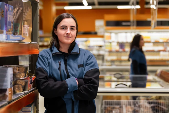 Ung kvinna jobbar i butik, står med armarna i kors och har ett bestämt uttryck.