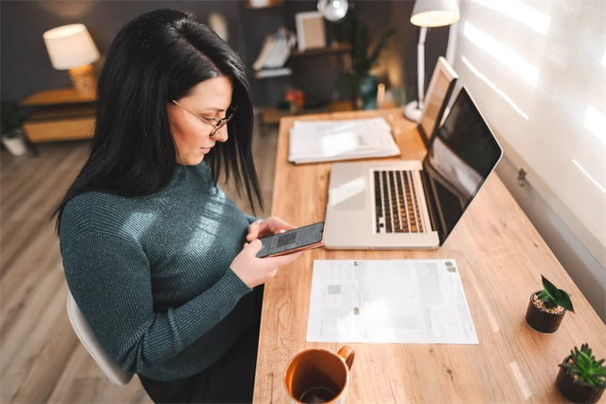 Kvinna sitter vid skrivbord med dator och mobiltelefon.